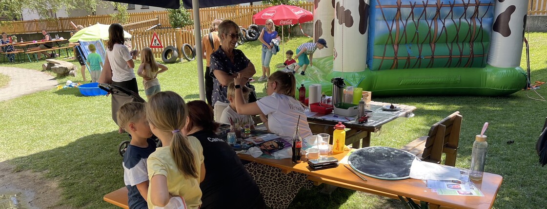 Kinder und Erwachsene stehen und sitzen unter einem Sonnenschirm in einem Garten. Im Hintergrund eine kleine Hüpfburg.