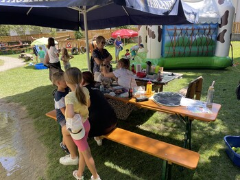 Kinder und Erwachsene stehen und sitzen unter einem Sonnenschirm in einem Garten. Im Hintergrund eine kleine Hüpfburg.