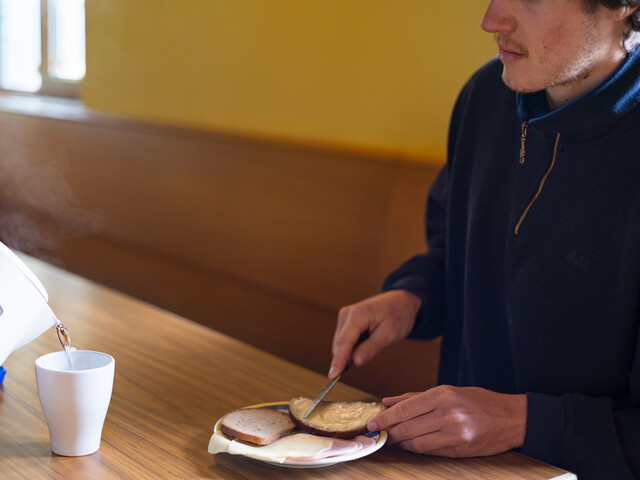 Eine Person sitzt an einem Tisch und streicht sich ein Butterbrot und bekommt eine Tasse heißen Tee eingeschenkt.