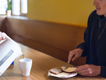 Eine Person sitzt an einem Tisch und streicht sich ein Butterbrot und bekommt eine Tasse heißen Tee eingeschenkt.