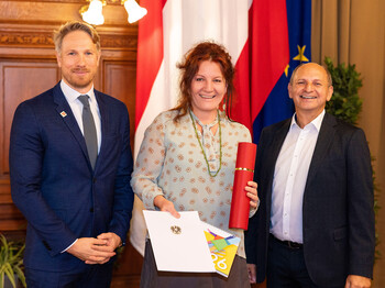 Eine Caritas Mitarbeiterin steht zwischen zwei Männern in Anzügen. Im Hintergrund sieht man die Flagge Österreichs und der Europäischen Union.
