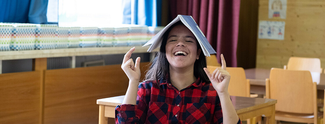 Eine Schülerin mit einem Übungsbuch auf dem Kopf, lacht in die Kamera.