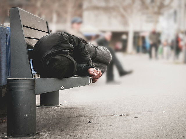 In einer Fußgängerzone schläft ein Mann mit Winterjacke und Mütze auf einer Bank.