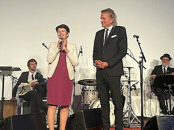 Zwei Personen stehen vor einer Band auf einer Bühne. Die Frau spricht in ein Mikrofon.