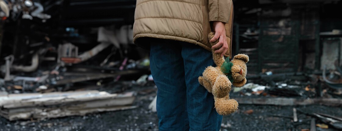 Ein Kind vor einem Haufen Trümmern hält einen Teddybären in der rechten Hand.