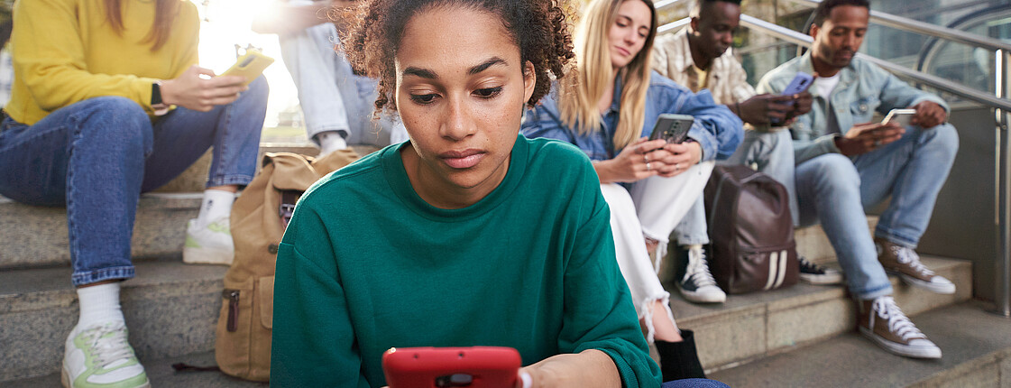 Sechs Jugendliche sitzen auf einer Betontreppe und schauen fokussiert auf ihre Smartphones.