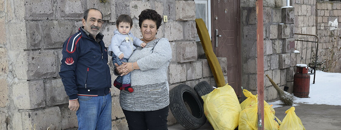 Eine Mutter mit Kind auf dem Arm und der Vater stehen vor einer Notunterkunft. Im Hintergrund liegen Säcke mit Hilfslieferungen auf dem Boden.