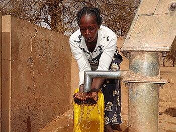 Eine Frau mit gelbem Wasserkanister steht an einer Wasserpumpe und hält ihre Hände unter das laufende Wasser.
