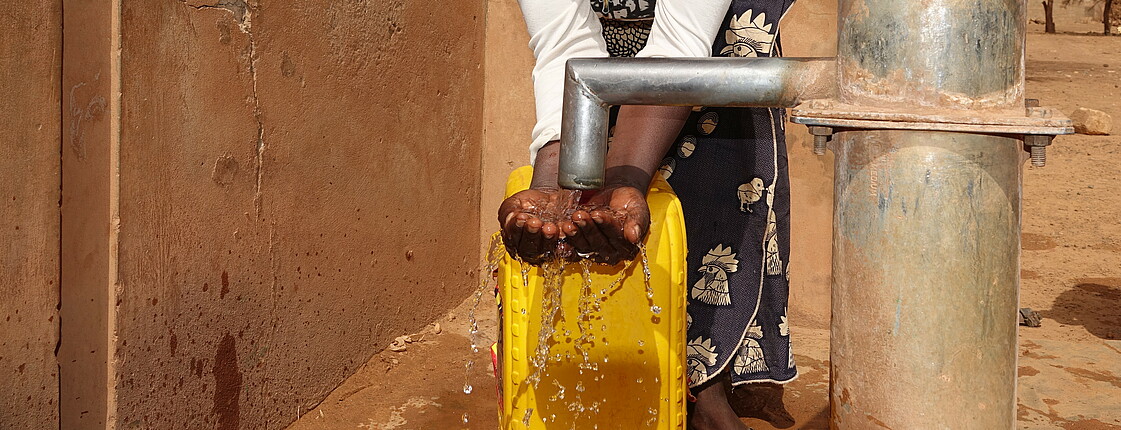 Eine Frau mit gelbem Wasserkanister steht an einer Wasserpumpe und hält ihre Hände unter das laufende Wasser.