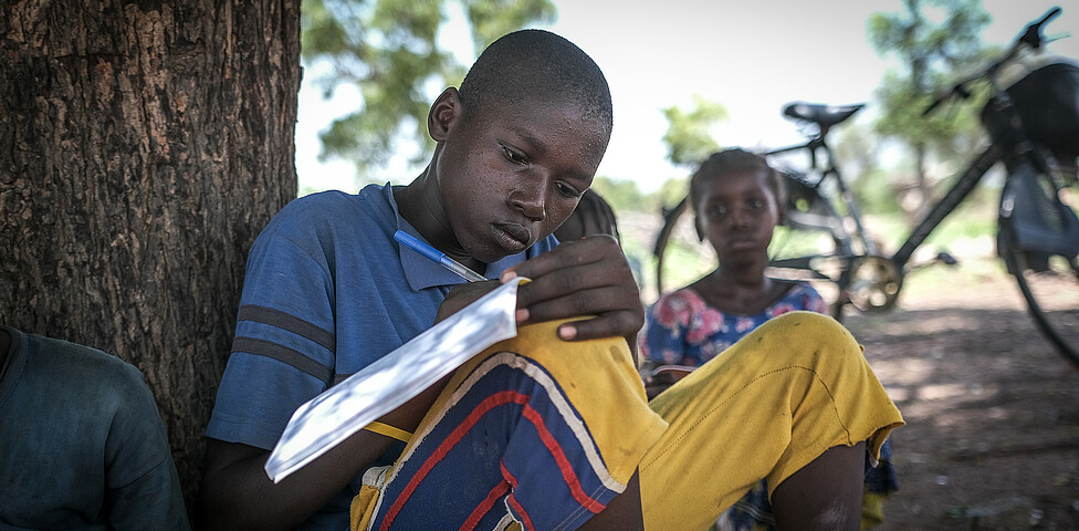 Ein afrikanischer Jugendlicher, der an einem Baum sitzt, schreibt in ein Heft.
