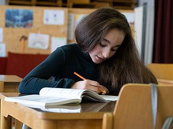 Ein Mädchen sitzt an einem Tisch und lehnt konzentriert über einem Schulaufgabenheft.