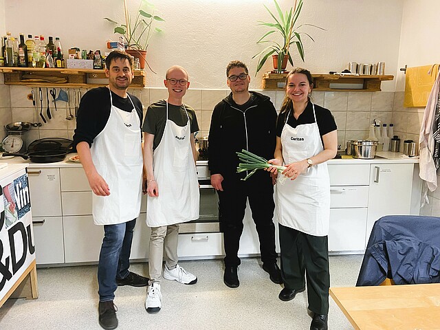 Vier Personen mit weißen Kochschürzen stehen nebeneinander in einer Küche.