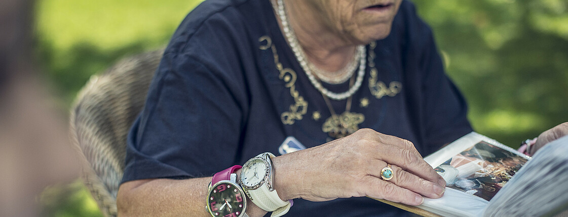 Eine ältere Frau mit Demenz blättert im Garten durch ein Fotoalbum. An ihrem rechten Arm trägt sie zwei Armbanduhren.