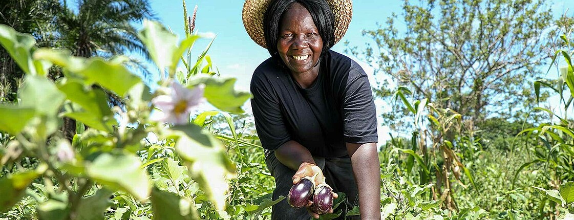Portrait einer lächelnden Frau mit Strohhut, die in einem Gemüsefeld steht und zwei Aubergine in die Kamera hält.