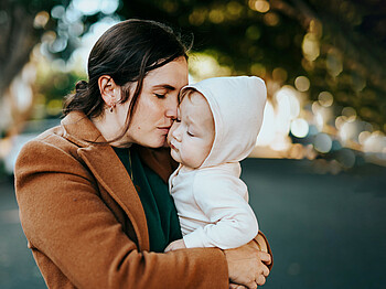 Eine Frau mit langem braunen Mantel drückt ein Kind in ihren Armen fest an sich.
