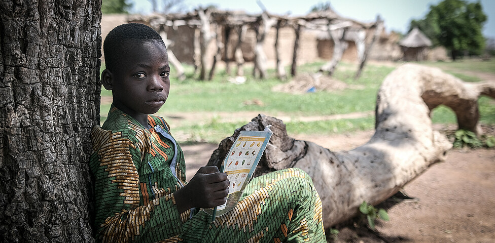Portrait eines afrikanischen Jungen, der an einem Baum lehnend sitzt, und in einem Heft liest.