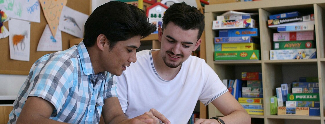 Zwei junge Männer sitzen an einem Tisch und schauen in ein Lernhilfebuch.