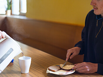 Eine Person sitzt an einem Tisch und streicht sich ein Butterbrot und bekommt eine Tasse heißen Tee eingeschenkt.