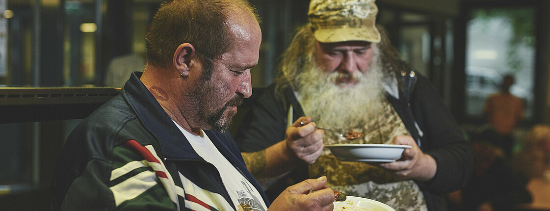 Zwei Männer ohne Obdach nehmen eine warme Mahlzeit zu sich.
