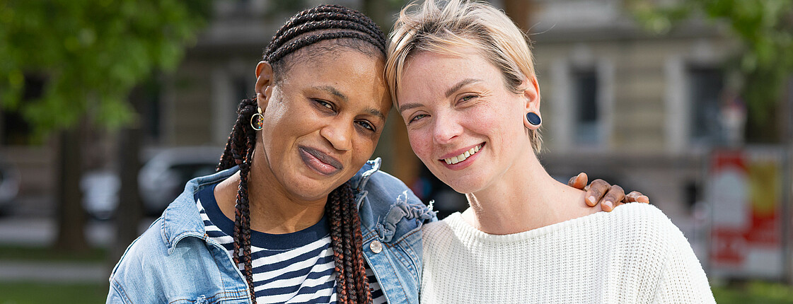 Zwei junge Frauen, die sich im Buddy-Projekt kennengelernt haben, stehen Arm in Arm in einem Park.