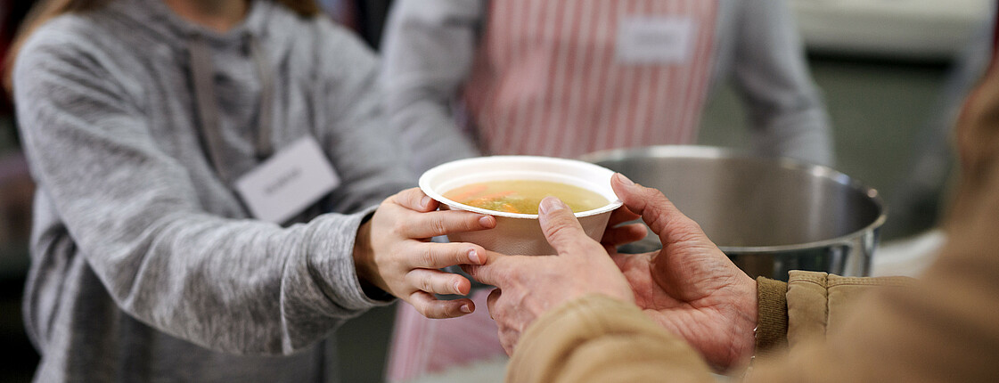 Eine Frau mit Mundschutz händigt einem Mann eine Schüssel mit warmer Suppe aus.