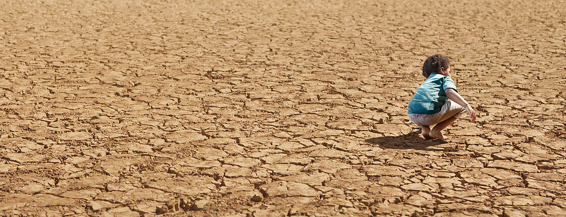 Ein kleines Kind hockt barfuß in einer ausgetrockneten Landschaft, welche bis zum Horizont von Dürre gezeichnet ist.