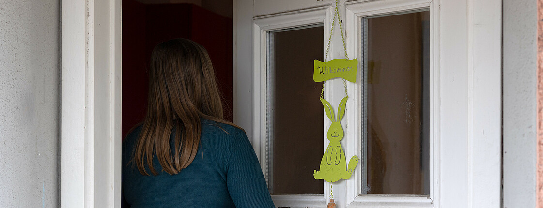 Eine junge Frau betritt durch eine weiße Haustür mit dem Schild "Willkommen" eine Einrichtung.