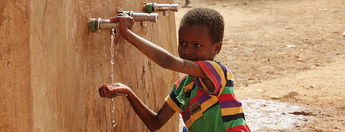 Ein Junge in bunt gestreiftem T-Shirt steht an einem Trinkbrunnen und lässt Wasser in seine rechte Hand tropfen.