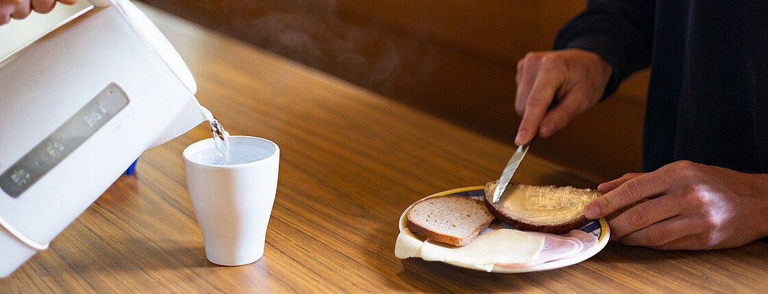 Eine Person sitzt an einem Tisch und streicht sich ein Butterbrot und bekommt eine Tasse heißen Tee eingeschenkt.